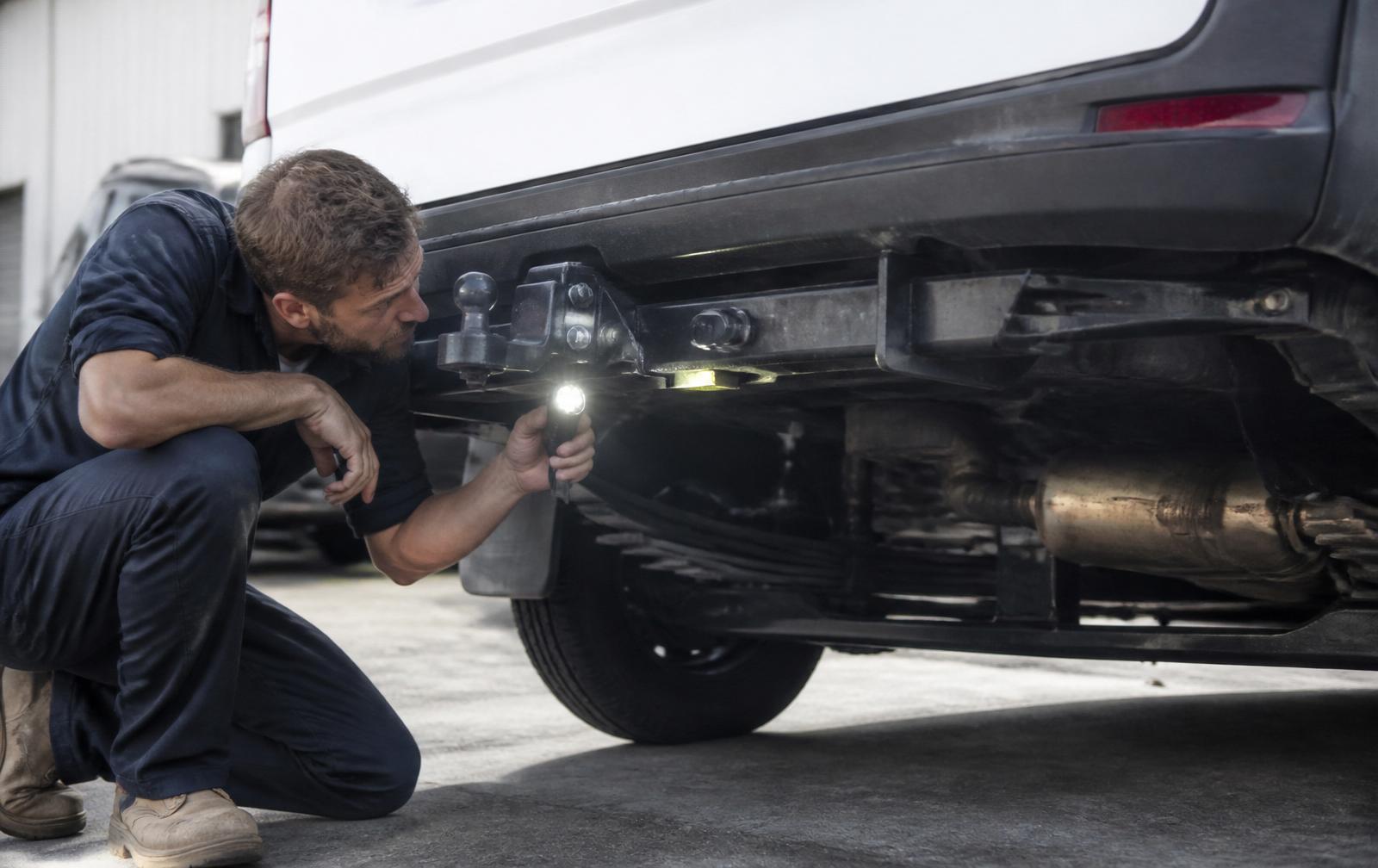 Mechanic in navy workwear crouched beneath the rear of a white van with an LED torch, inspecting the tow bar mount, leaf spring suspension and chassis rail